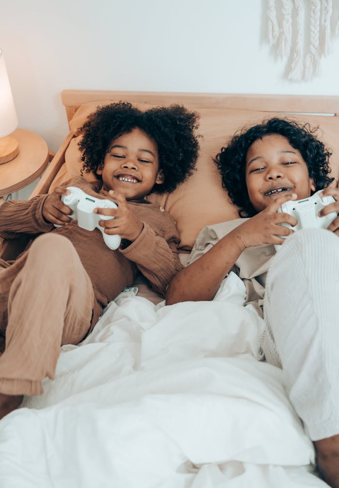 From above of happy African American sisters in domestic wear lying on comfortable bed together and playing video game with gamepads during free time at home